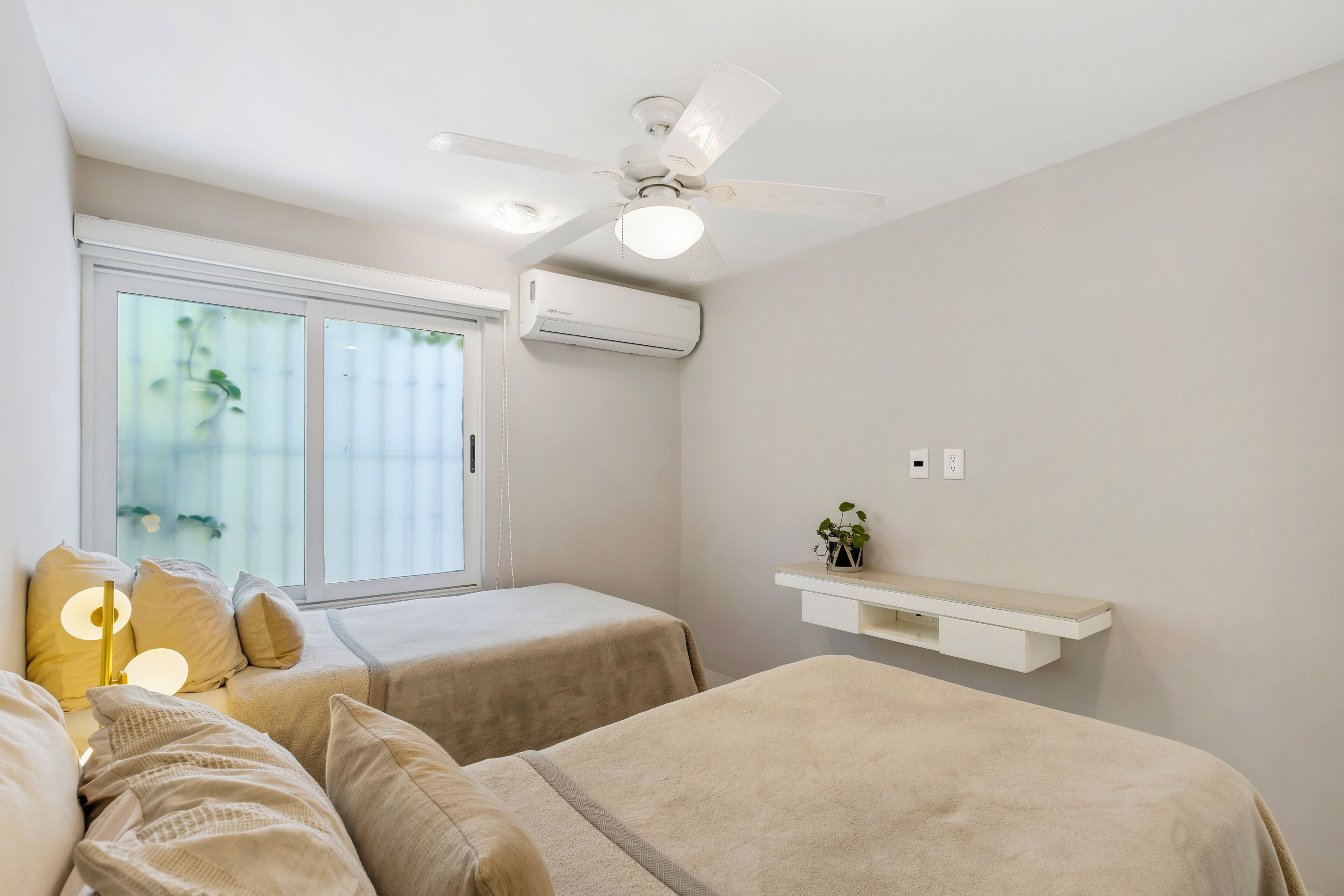 A bedroom with two beds, a ceiling fan, an air conditioning unit, a window with blinds, and a bedside table with a plant.