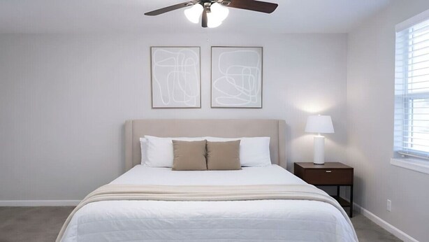 Un dormitorio con cama, ventilador de techo, mesita de noche con lámpara y arte abstracto en la pared.