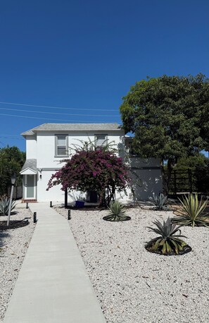 A white house with a concrete pathway leading to the entrance, surrounded by various plants and a large tree. - Antique Row Retreat (West Palm Beach)