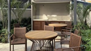En uteplats med en träpergola, runda bord, flätade stolar och en inbyggd grill.