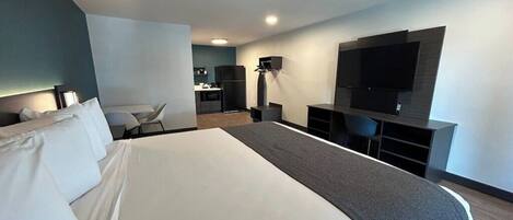 Et moderne hotelværelse med en stor seng, et skrivebord, en stol, et fladskærms-tv monteret på væggen og et tekøkken i baggrunden.