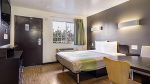 Ein Hotelzimmer mit Bett, Schreibtisch, Stuhl, einem Fenster mit Vorhängen und einer Wandleuchte.