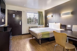 Habitación de hotel con cama, escritorio, silla, ventana con cortinas y lámpara fijada a la pared.