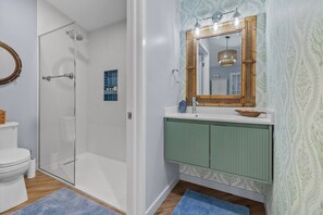 Ein Badezimmer mit Duschkabine aus Glas, einer weißen Toilette, einem grünen Waschtisch mit Spiegel und Wandpapier mit Muster.