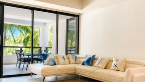 A modern living room with a beige sectional sofa, a dining table with chairs, and a large sliding glass door leading to an outdoor area with palm trees.
