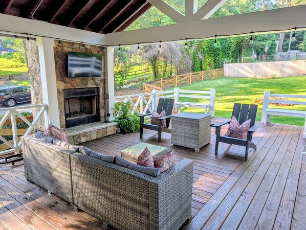 A covered outdoor seating area with wicker furniture, a stone fireplace, and a view of a green backyard.