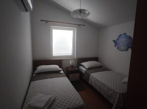 A small, dimly lit room with two single beds, a window with blinds, a wall-mounted lamp, and a small table with a lamp. - Two Bedroom Apartment with Balcony and Sea View (Karlobag)