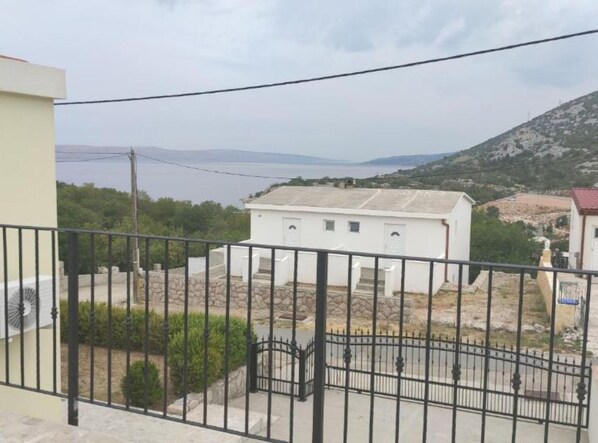 A white building with a small porch, a black metal fence, and a view of a hilly landscape with a body of water in the distance. - Two Bedroom Apartment with Balcony and Sea View (Karlobag)