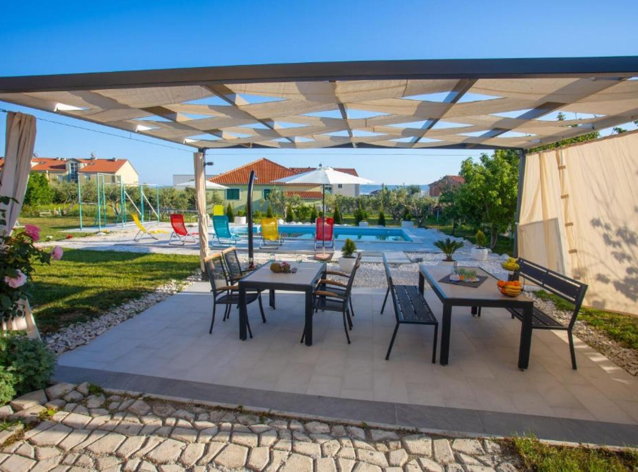Un porche cubierto con mobiliario exterior, una piscina y vistas a casas al fondo.
