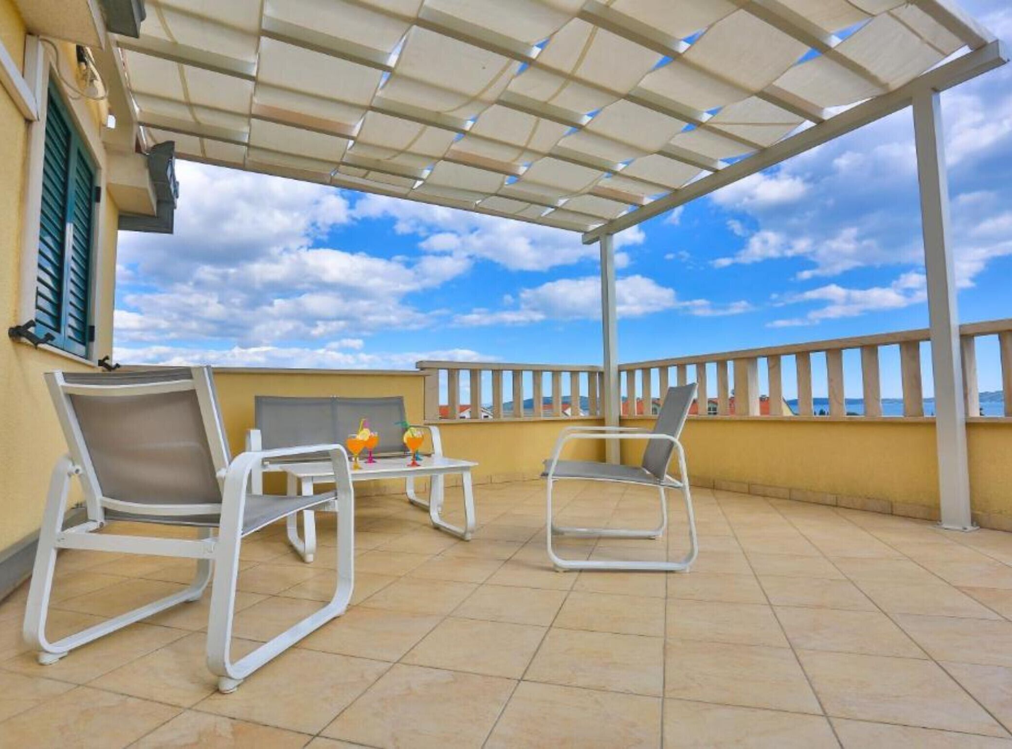 Un'area salotto esterna coperta con due sedie e un tavolo, vista sul mare e un edificio con facciata gialla.