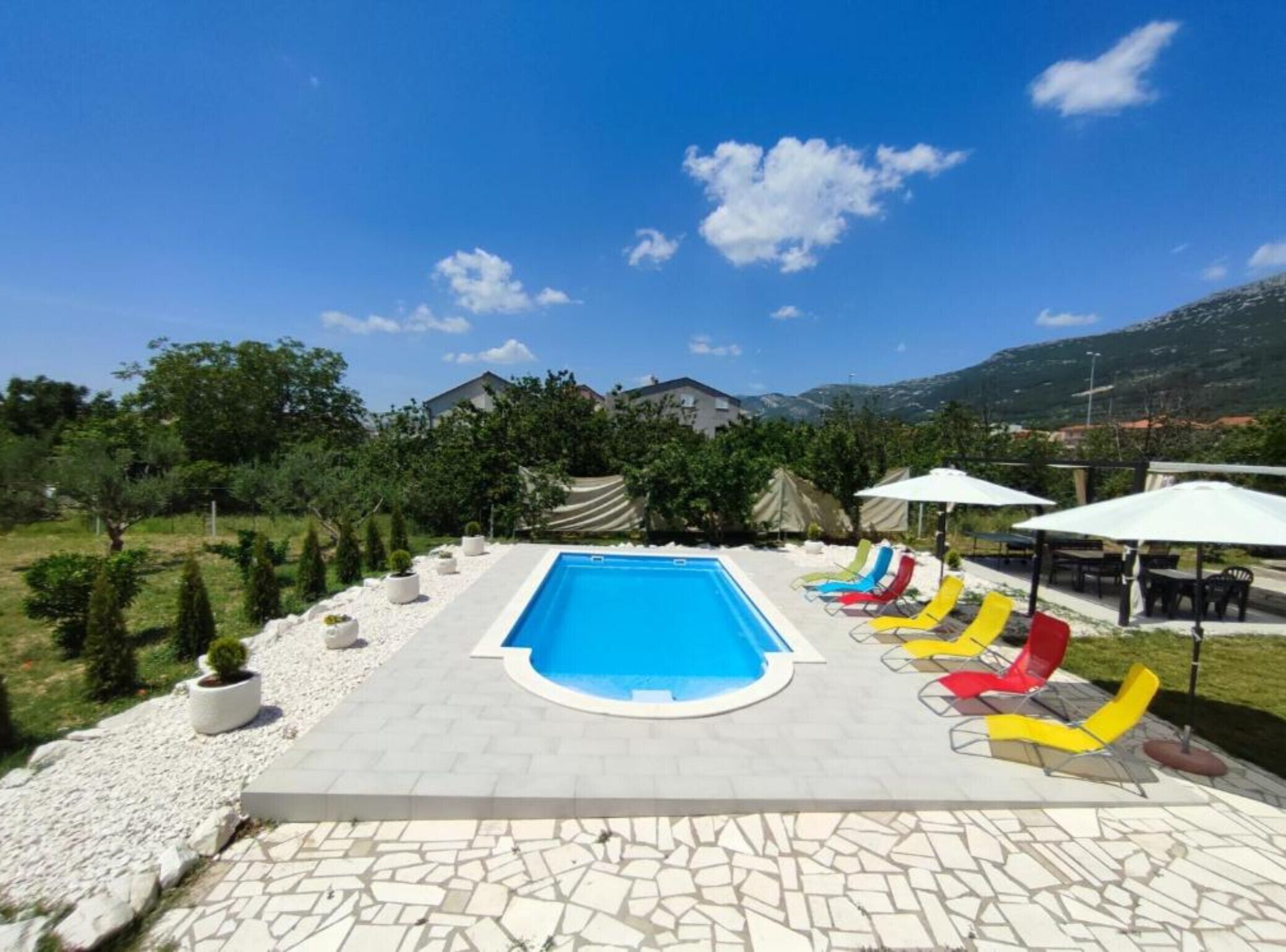 Une piscine entourée de chaises longues et de parasols, avec une végétation luxuriante et des bâtiments en arrière-plan.