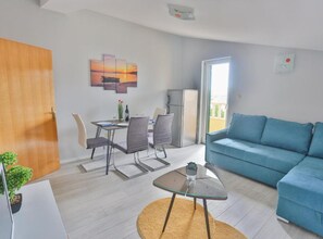 Ein modernes Wohnzimmer mit einem blauen Sofa, einer Essecke mit Tisch und Stühlen, einem Bild an der Wand und Blick durch eine Tür.