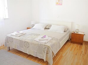 Ein ordentlich bezogenes Bett mit einer gemusterten Tagesdecke, zwei Kissen und zwei gefalteten Handtüchern auf der Seite. Ein Nachttisch mit einer Lampe und einem kleinen Dekorationsgegenstand steht daneben. Der Raum verfügt über Holzböden und eine schlichte weiße Wand.