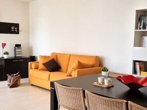 Ein modernes Wohnzimmer mit einer Couch, einem Esstisch, Korbstühlen, einem Bücherregal und einer kleinen Pflanze.