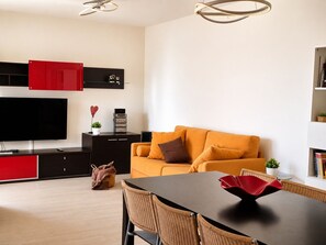 Ein modernes Wohnzimmer mit einer Couch, einem Esstisch, einem Fernseher und einer Küchenzeile mit Schränken und einer roten Tür.