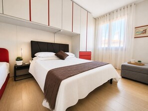 Ein Hotelzimmer mit Bett, Nachttischen, einem roten Sessel, einem grauen Sofa und einem Fenster mit Vorhängen.