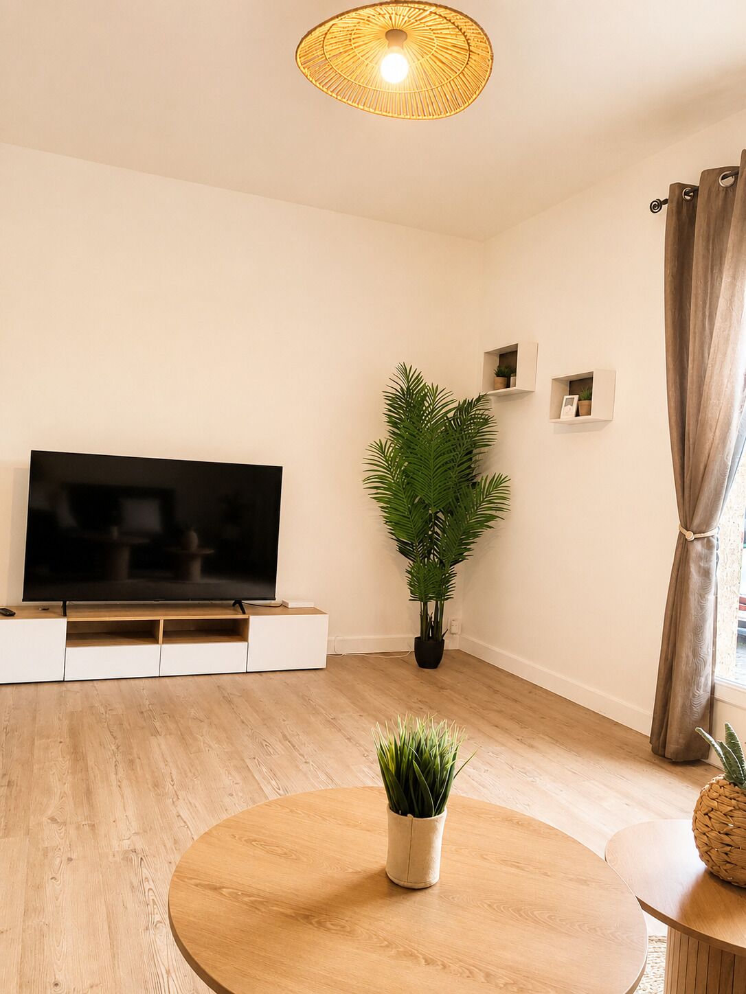 Un salon équipé d’un téléviseur à écran plat, d’une table basse ronde en bois, d’une plante en pot et d’un luminaire fixé au plafond.