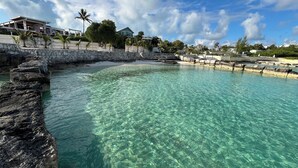 Una piscina turquesa y transparente rodeada de un muro de piedra, con una playa y casas al fondo.