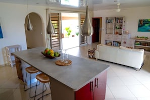 A modern kitchen with a central island, bar stools, a dining table, and a living area with a sofa and bookshelves. - Dolce Vita Villa Moorea (Moorea-Maiao)