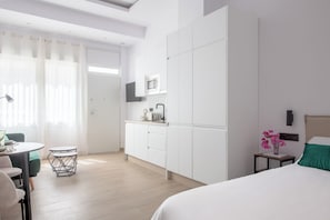 A modern hotel room with a bed, a small kitchenette, a dining area with a table and chairs, and a large window with curtains.