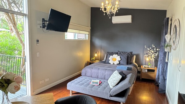 A bedroom with a bed, a TV mounted on the wall, a chandelier, an air conditioner, and a window with a view of greenery.