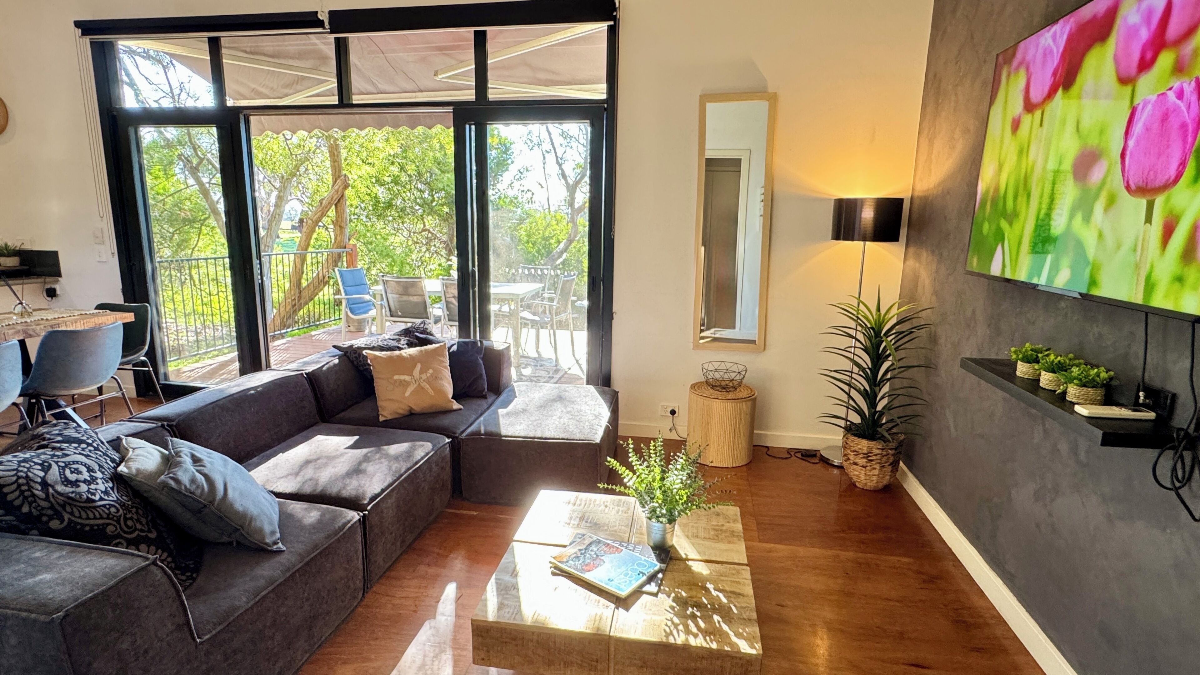 A modern living room with a sectional sofa, a coffee table, a TV mounted on the wall, and a view of the outdoors through large windows.