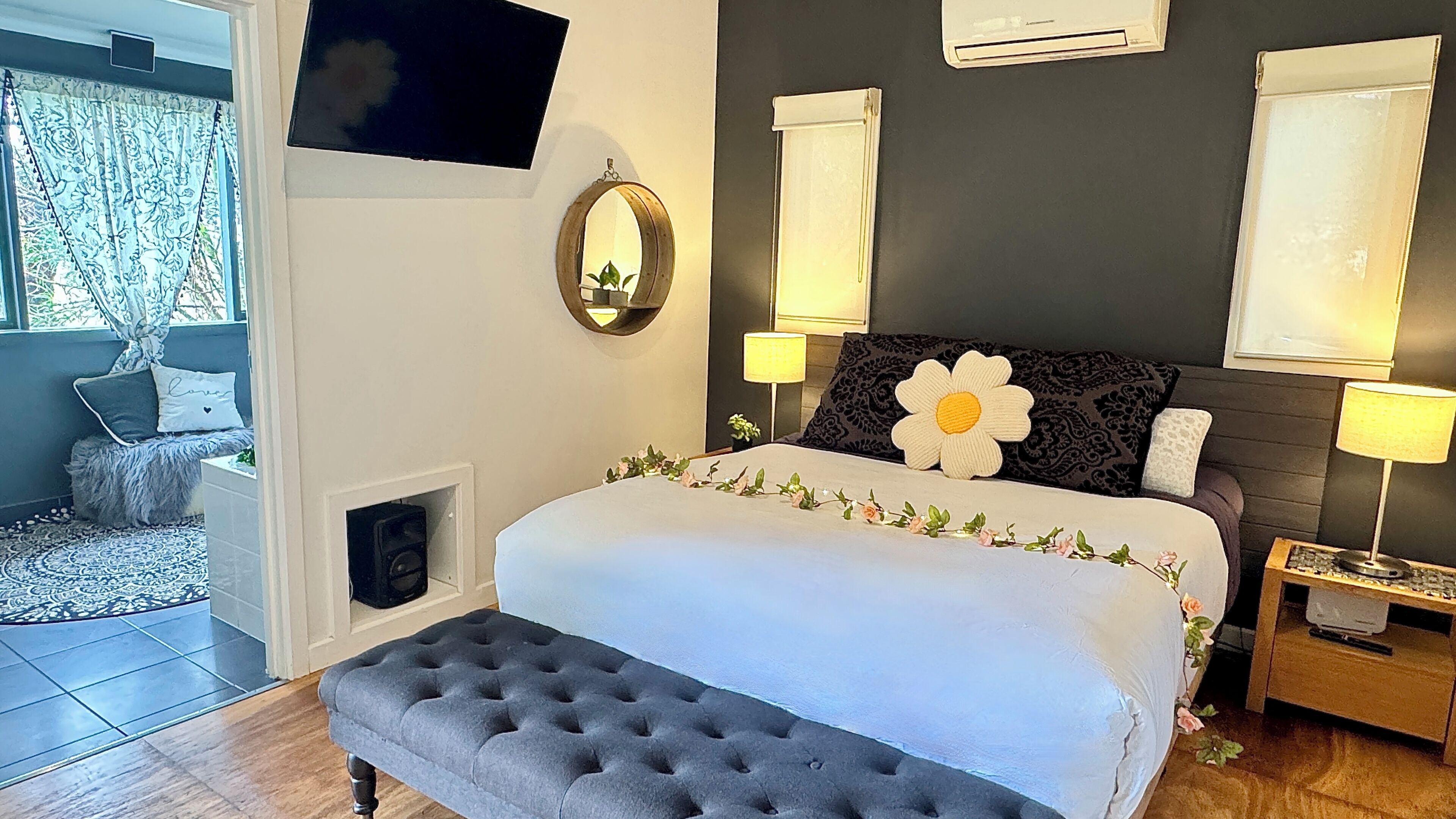 Un dormitorio con cama, lámparas de noche, televisión, ventana con cortinas y un pequeño banco.
