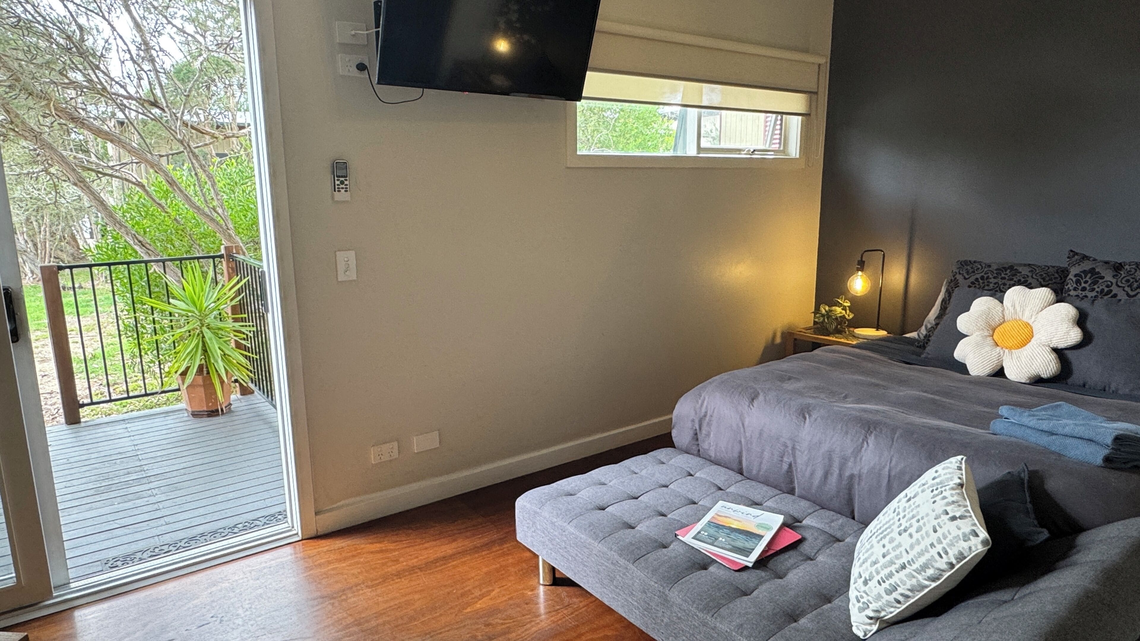 A bedroom with a grey sofa, a bed with a white flower pillow, a nightstand with a lamp, and a TV mounted on the wall.