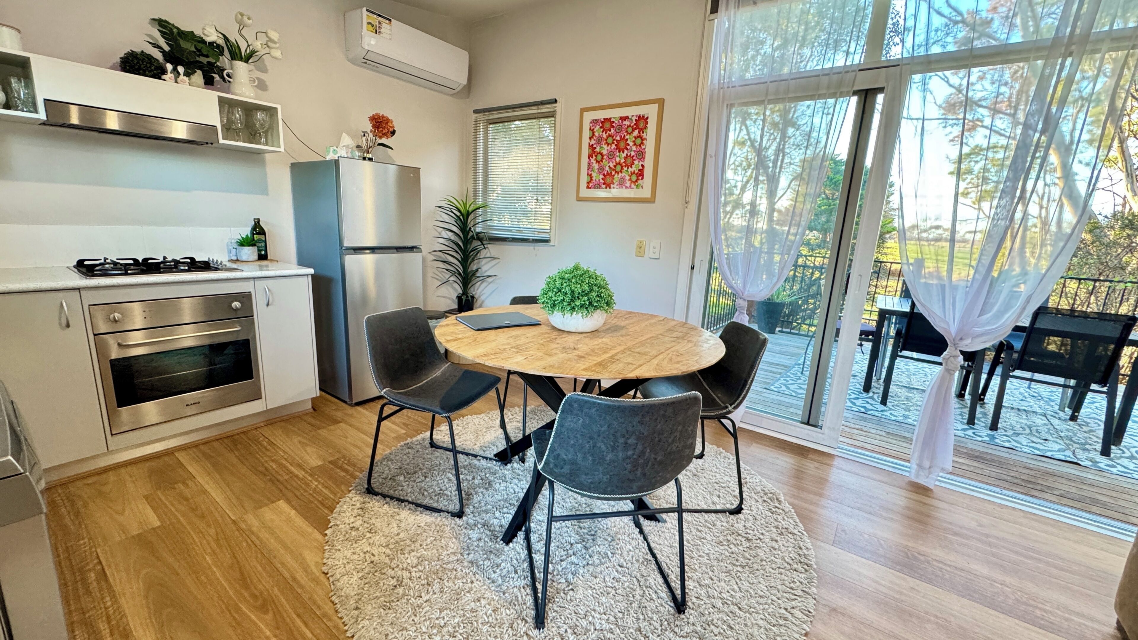 Una cocina moderna con estufa a gas, horno y refrigerador, un área de comedor con una mesa redonda y sillas, y un ventanal corredero que da a un patio exterior.