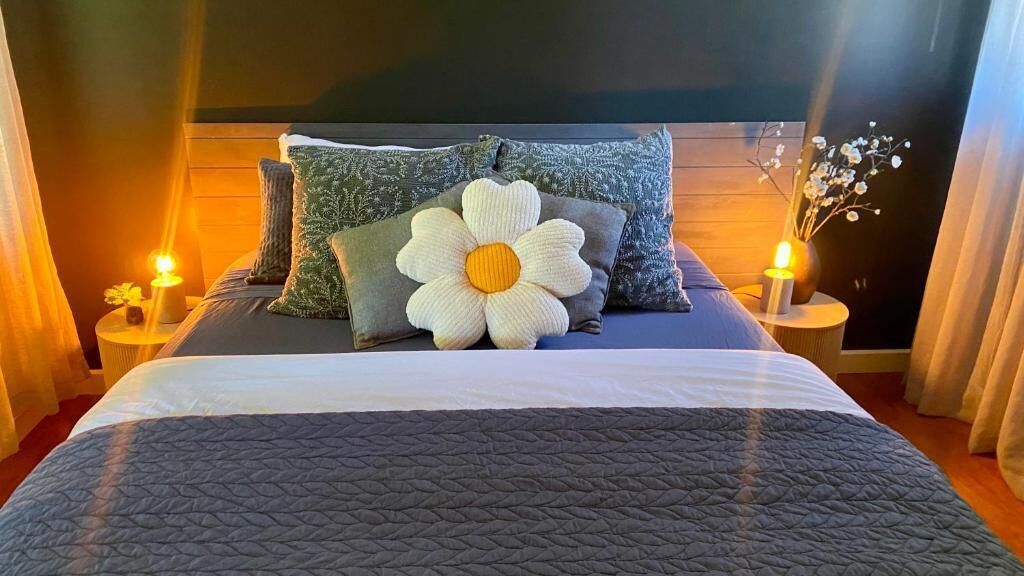 Ein ordentlich bezogenes Bett mit einem großen Blumenkissen, zwei Nachttischen mit Kerzen und einem dunklen Kopfteil.