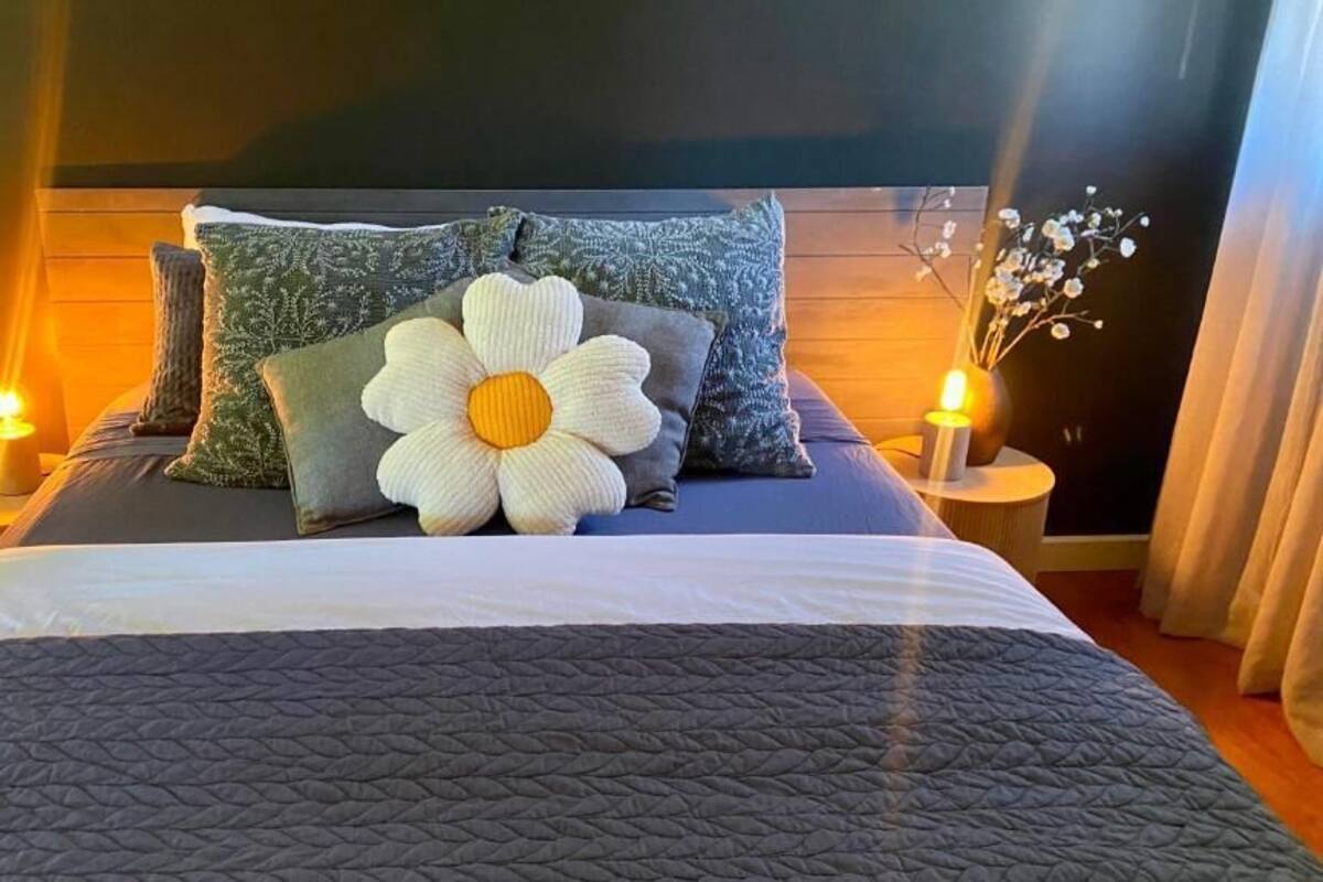 Una cama bien hecha con un almohadón grande de flores, dos mesitas de noche con velas y un cabecero oscuro.