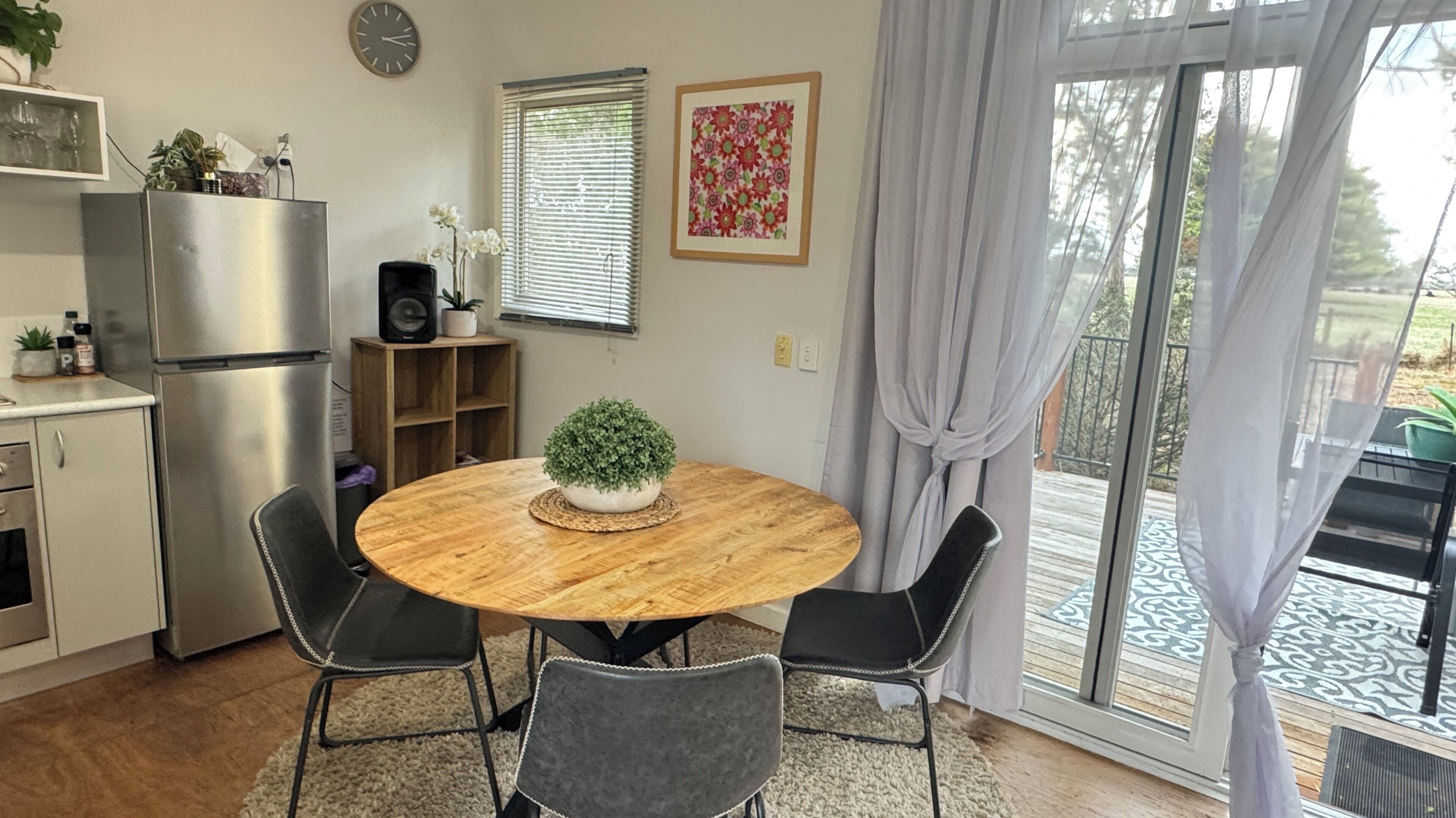 Una cocina compacta con refrigerador, una mesa de madera con cuatro sillas, un estante con plantas y una ventana con persianas.