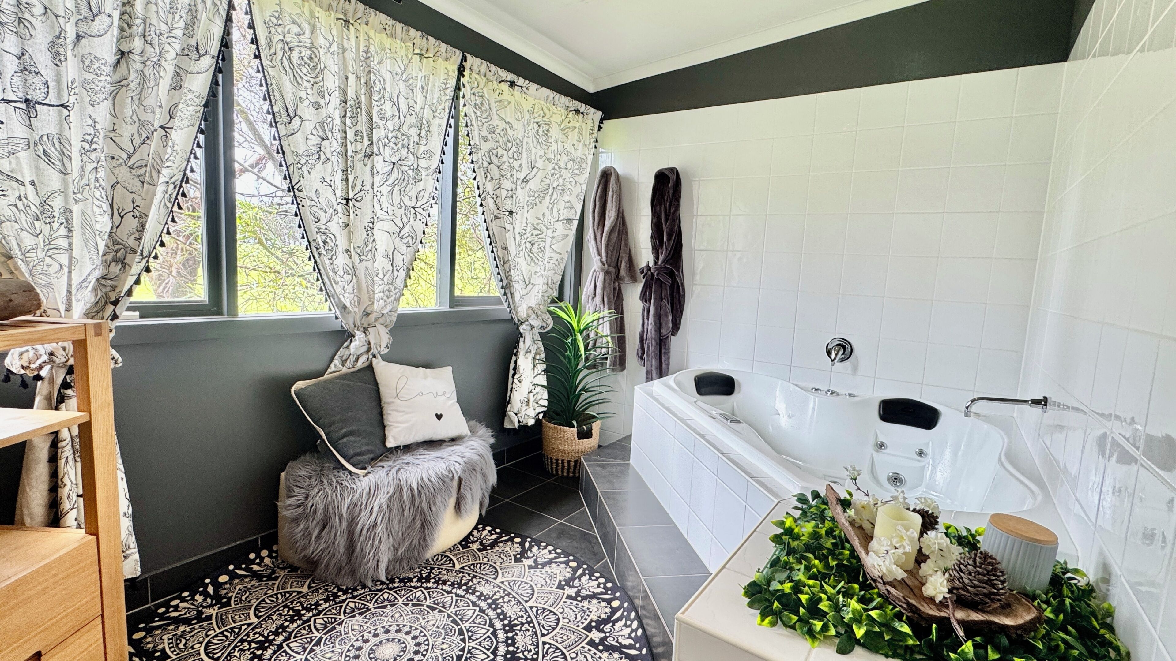 A modern bathroom with a bathtub, a window with floral curtains, a fluffy stool, and a decorative plant arrangement.