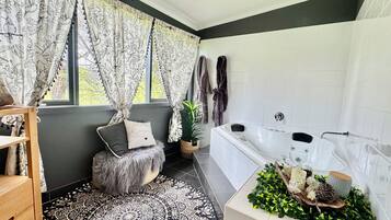 A modern bathroom with a bathtub, a window with floral curtains, a fluffy stool, and a decorative plant arrangement.