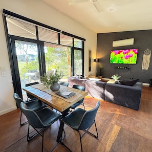A modern living room with a dining area, large windows, a flat-screen TV, and a grey sofa.