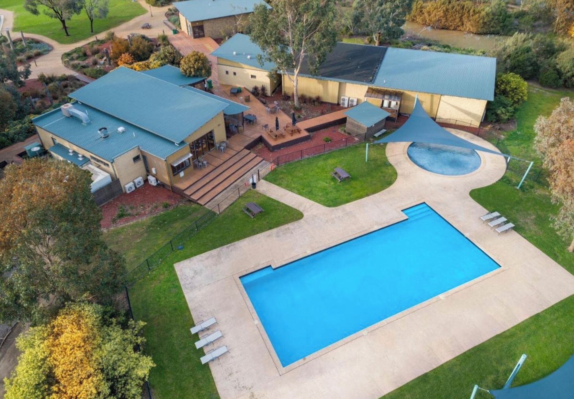 A large swimming pool with a surrounding deck, a covered patio area, and a building with a green roof.