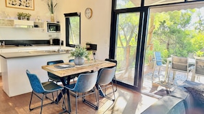 Una cocina moderna con microondas integrado, un comedor con mesa de madera y sillas azules, y un amplio ventanal corredera que da a un patio exterior.