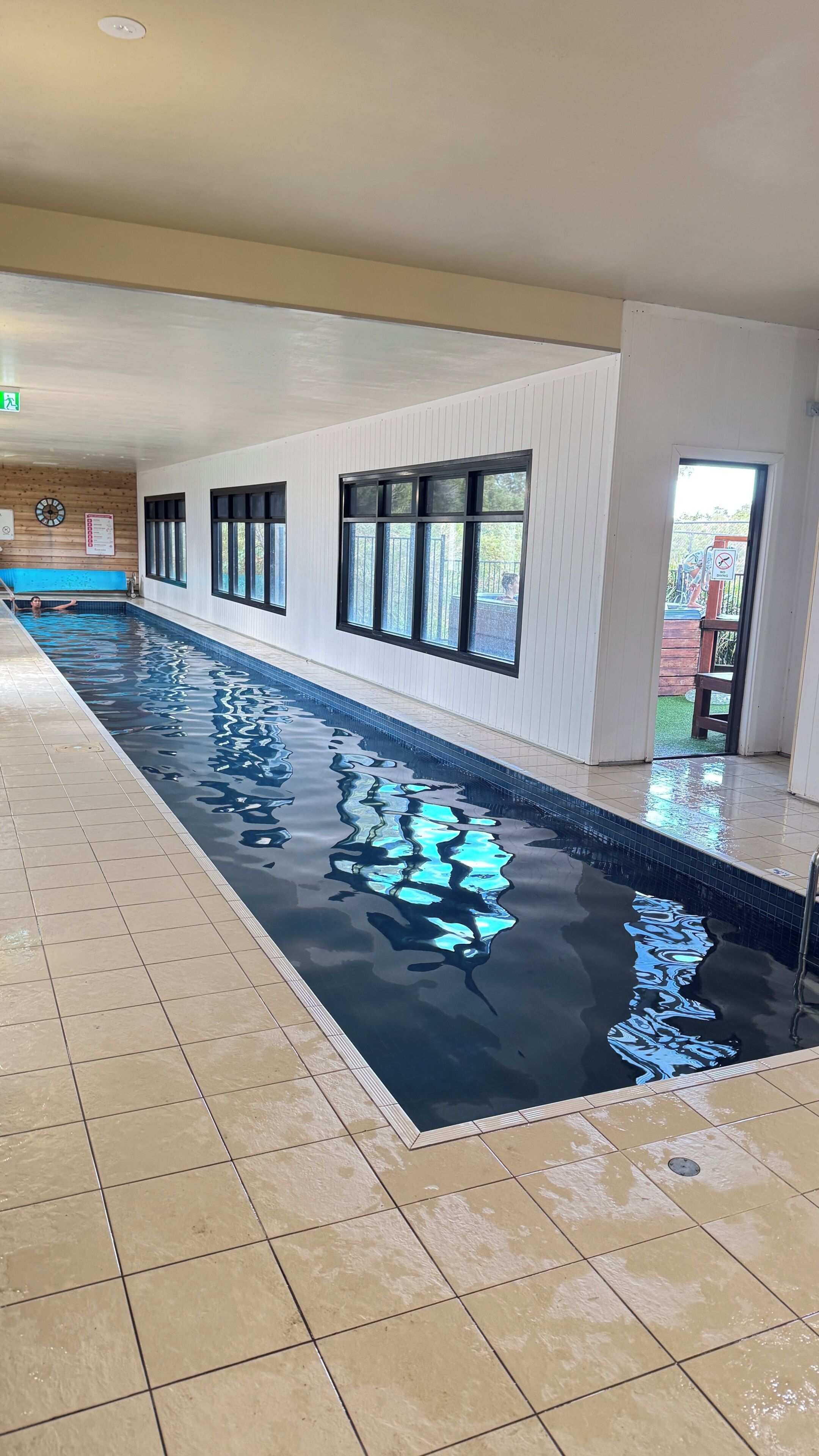 A long indoor swimming pool with clear water, tiled floor, and large windows.