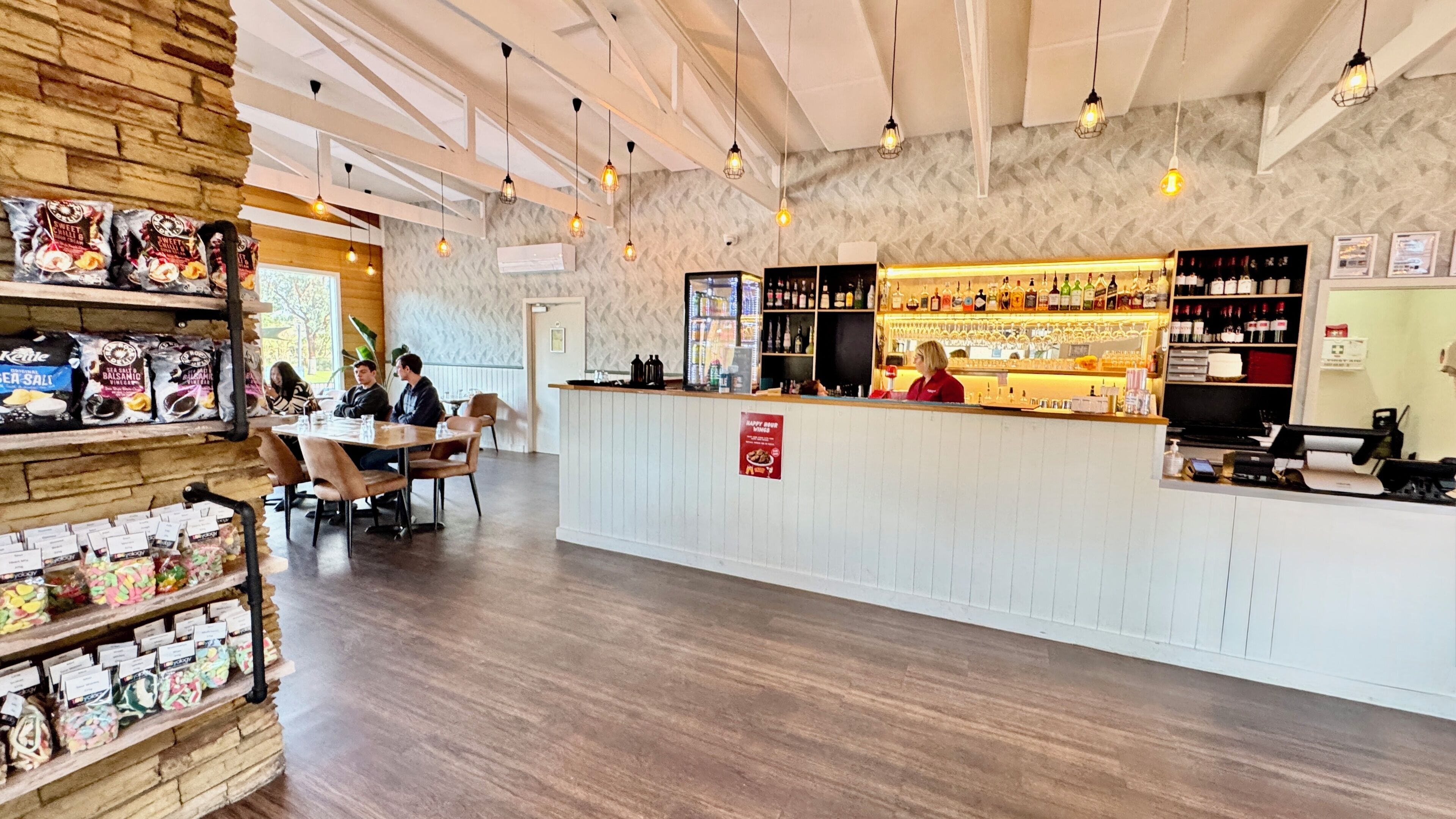 Un restaurante con área de bar, estantes de madera con aperitivos y una barra donde una persona está atendiendo.