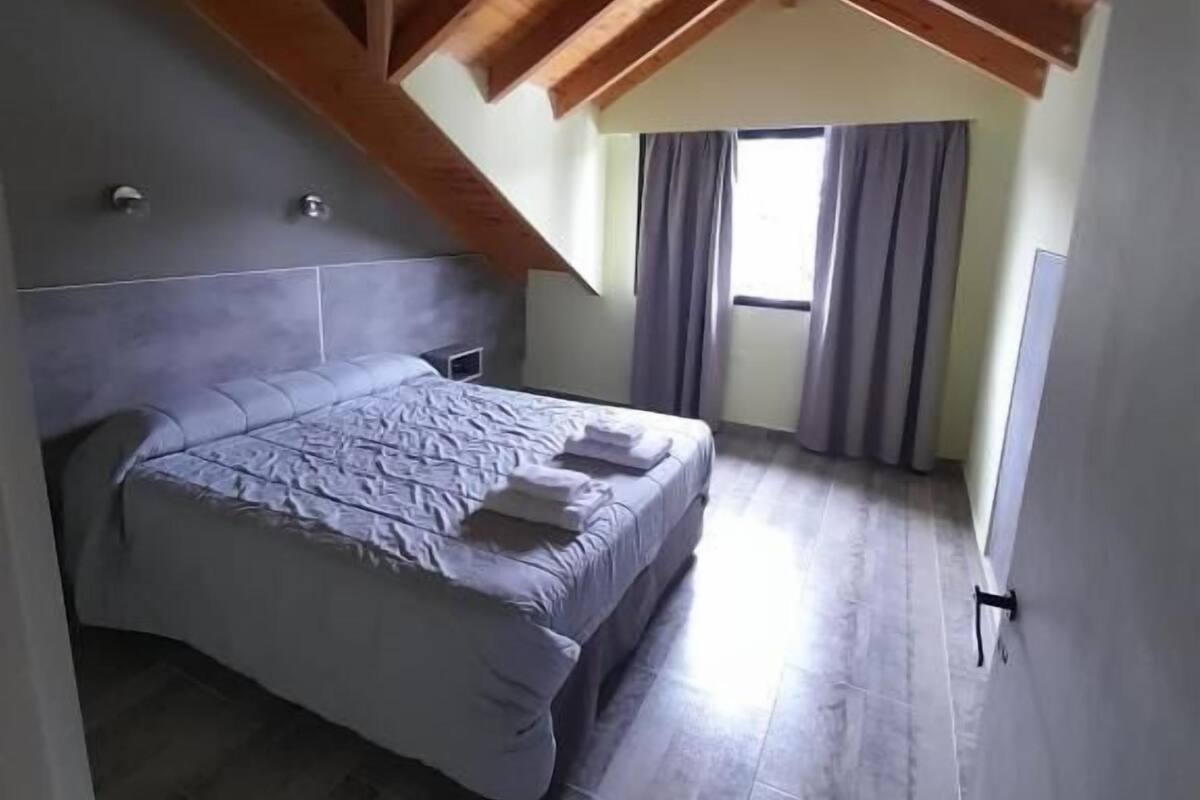 A bedroom with a bed, wooden ceiling beams, a window with curtains, and a tiled floor.