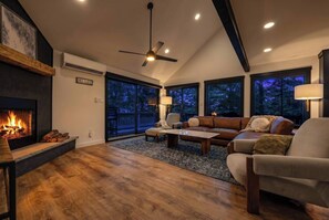 A living room with a fireplace, a ceiling fan, and a large window overlooking a deck. - Designer Ski In/Out Retreat | Pool, Spa & More (Tannersville)
