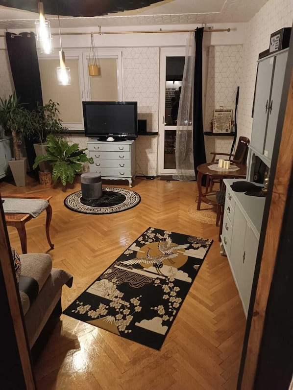 Una sala de estar con televisor, piso de madera, alfombra blanco y negro, y un comedor con mesa y sillas.