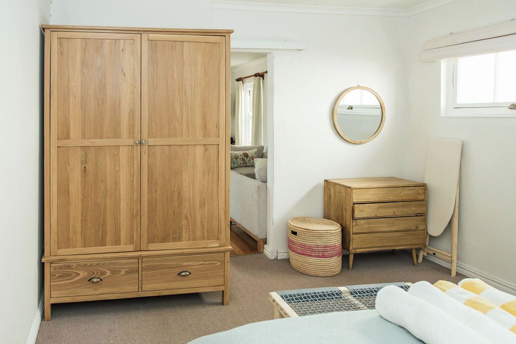 Une chambre avec une armoire en bois, une petite commode en bois, un miroir rond, un lit recouvert d’une couvre-lit à motifs et une fenêtre avec des rideaux.