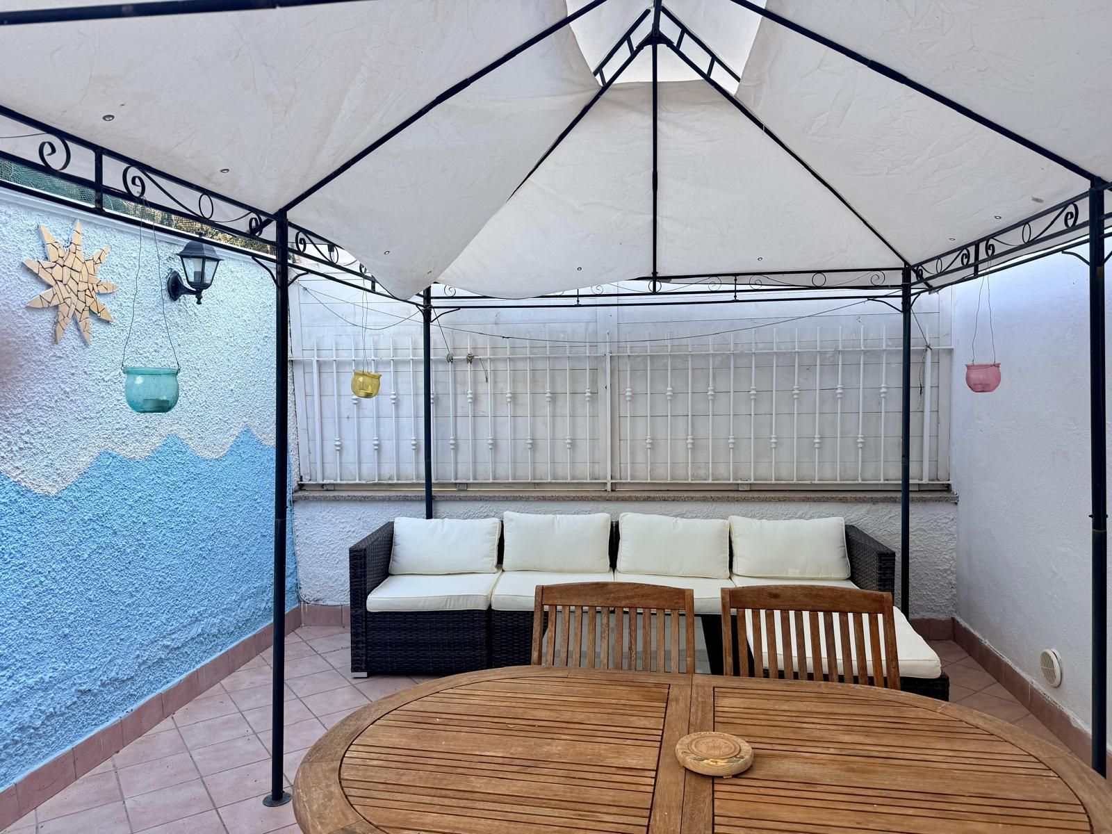 A covered patio area with a round wooden table, two sofas with white cushions, and a decorative wall with a sun motif.