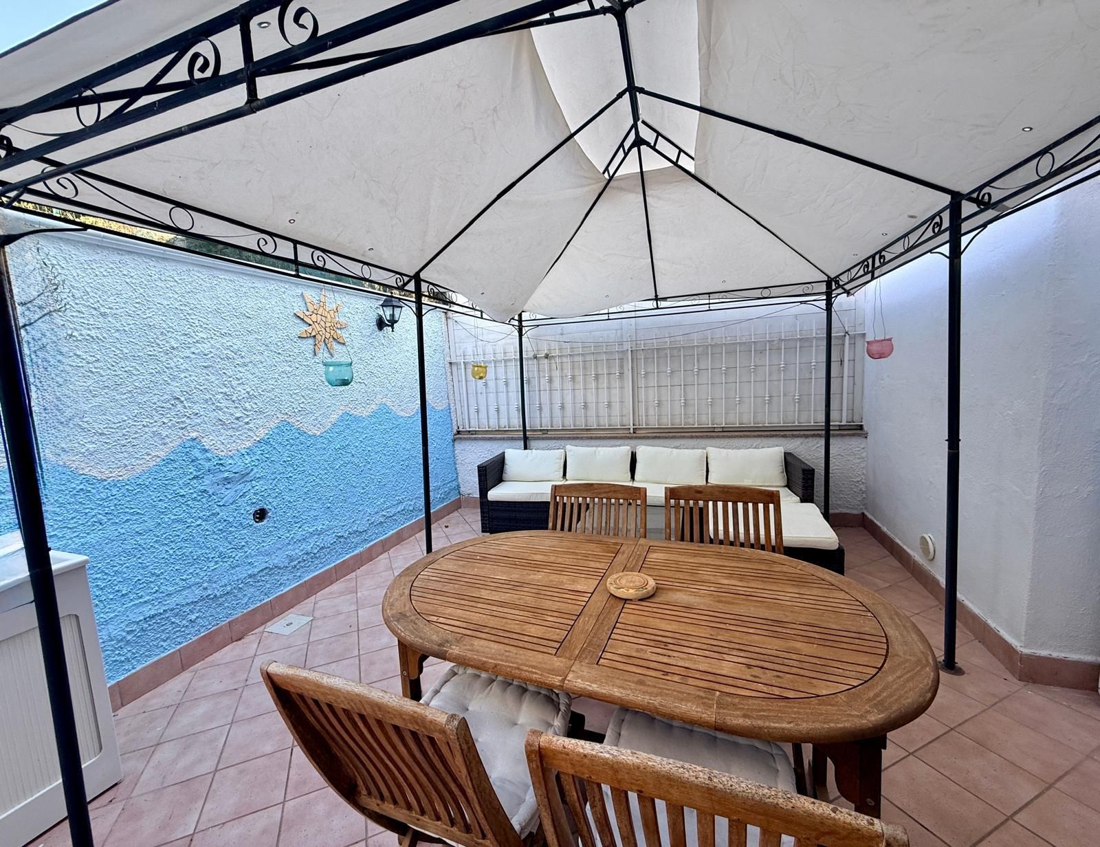 A covered patio area with a round wooden table, four chairs, a sofa, and a decorative wall.