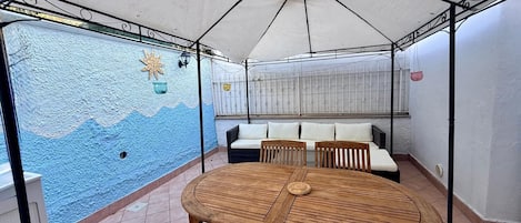 A covered patio area with a round wooden table, four chairs, a sofa, and a decorative wall.