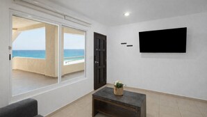 A room with a large window offering a view of the beach, a flat-screen TV mounted on the wall, and a dark wooden coffee table with a plant. - Large Group Getaway - Brisas Cancun Condo 3401 (Cancún)