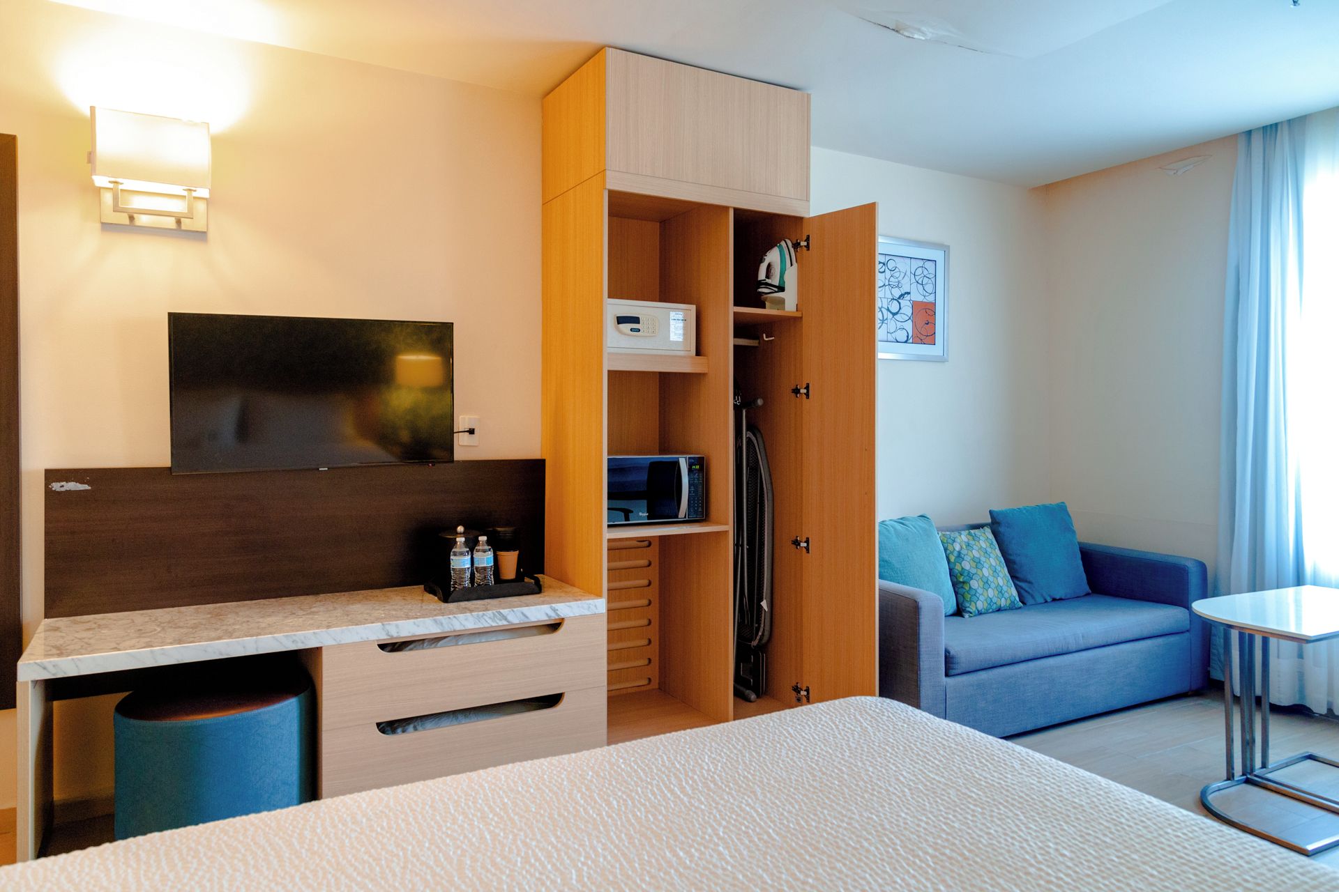 A hotel room with a bed, a desk with drawers, a microwave, a television, a sofa, and a small table.