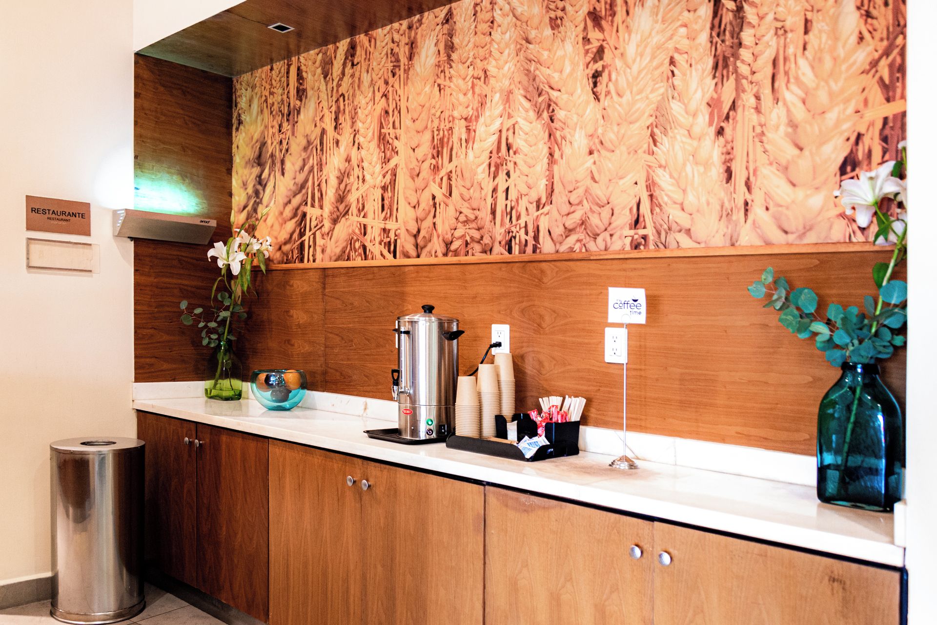 Zona de recepción de un hotel con mostrador de madera, estación de café y un cubo de basura.