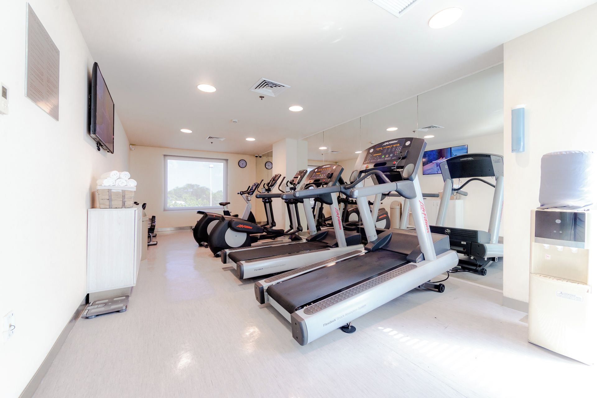 A gym with multiple treadmills, a wall-mounted TV, and a window providing natural light.